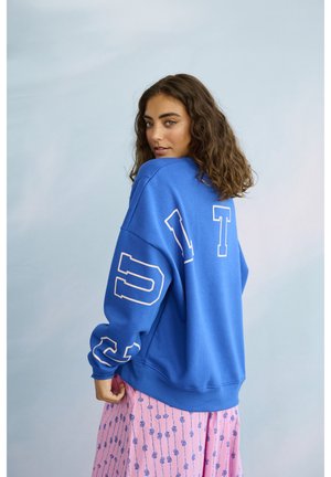 Žena s kučeravými vlasmi, oblečená v modrej mikine s bielymi písmenami a ružovej sukni so vzorom, pozerajúca cez rameno.