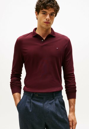 Polo à manches longues bordeaux avec une patte de boutonnage à trois boutons, orné d'un petit logo et associé à un pantalon texturé foncé.