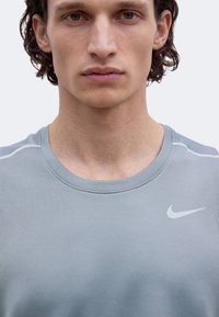 Jeune homme aux cheveux bouclés portant un débardeur de sport Nike gris clair sur un fond uni.