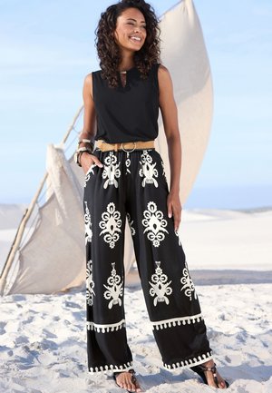 Schwarze weit geschnittene Hose mit weißen verschnörkelten Mustern und Quastenakzenten, kombiniert mit einem ärmellosen schwarzen Oberteil und einem geflochtenen Gürtel.