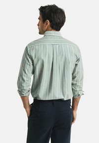 Camicia a righe verdi con maniche lunghe, polsini arrotolati e una piega sul retro, abbinata a pantaloni blu scuro.