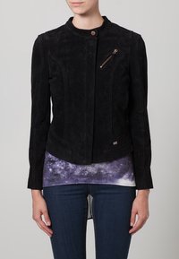 Zwarte suèden jas met een hoge kraag, aansluitend silhouet, een borstzak met rits en tonale stiknaden. Draagt een top met een galaxypatroon eronder.