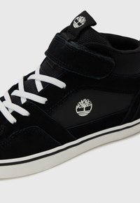 Black suede high-top sneaker with white laces, a velcro strap, and a logo on the side. Features a textured upper and rubber sole.