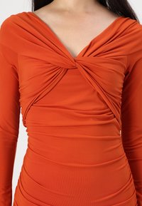 Haljina rust-orange boje s dugim rukavima, dubokim V-izrezom i skupljenim detaljima na prednjem dijelu, koji stvaraju drapirani efekt.