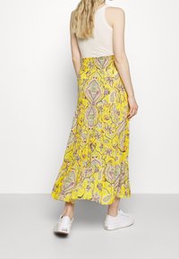 Larga falda amarilla con intrincado patrón de paisley en rosa, verde y púrpura. Tela fluida, cinturilla elástica y bajo en capas. Combinada con zapatillas blancas.