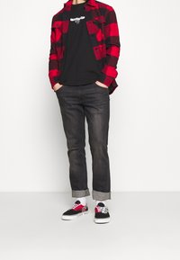 Camicia di flanella a quadri rossi e neri indossata sopra una t-shirt nera, jeans scuri e sneakers nere con accenti rossi e motivo a scacchi.