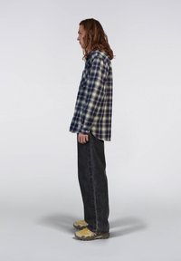 Camicia flanella a scacchi navy e crema, abbinata a pantaloni in denim neri e sneakers gialli. Mostra una vestibilità rilassata e maniche lunghe.