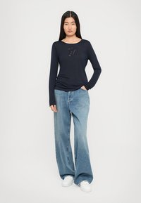 Žena stoji u tamnoplavoj majici dugih rukava s prozirnim dizajnom i širokima plavim trapericama, uz bijele tenisice.