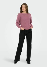 Femme debout, les mains dans les poches, portant un pull en tricot rose avec des boutons sur l'épaule et un pantalon noir droit.