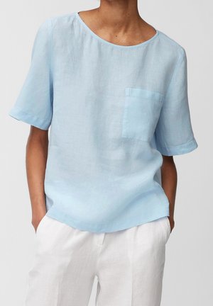 Kobieta ubrana w jasnoniebieską lnianą bluzkę z krótkim rękawem i przednią kieszenią oraz białe spodnie, z rękami w kieszeniach na jednolitym tle.