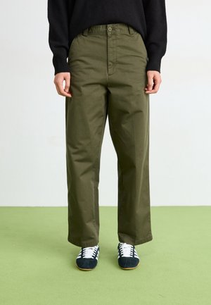 Personne portant un pantalon droit vert olive, un pull noir et des baskets noir et blanc, debout sur un sol vert.