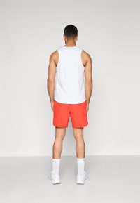 Λευκό αμάνικο πουκάμισο σε συνδυασμό με φωτεινά κόκκινα σορτς. Τα σορτς έχουν λεία υφή, και η στολή περιλαμβάνει λευκές αθλητικές κάλτσες και παπούτσια.