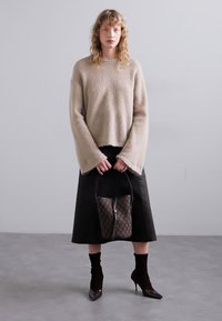 Beżowy sweter pleciony z szerokimi rękawami, czarna spódnica midi, teksturowane rajstopy oraz czółenka na obcasie z lakierowanej skóry, trzymając torebkę z monogramem.
