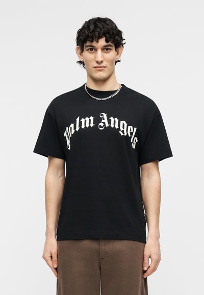 Νεαρός με σκούρα, σγουρά μαλλιά φοράει μαύρο t-shirt Palm Angels και καφέ παντελόνι, στέκεται μπροστά σε απλό φωτεινό φόντο.