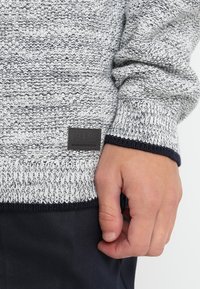 Maglione grigio e bianco lavorato a maglia con un motivo texturizzato, dotato di un polsino blu navy e un'etichetta nera con "1998" sull'orlo.