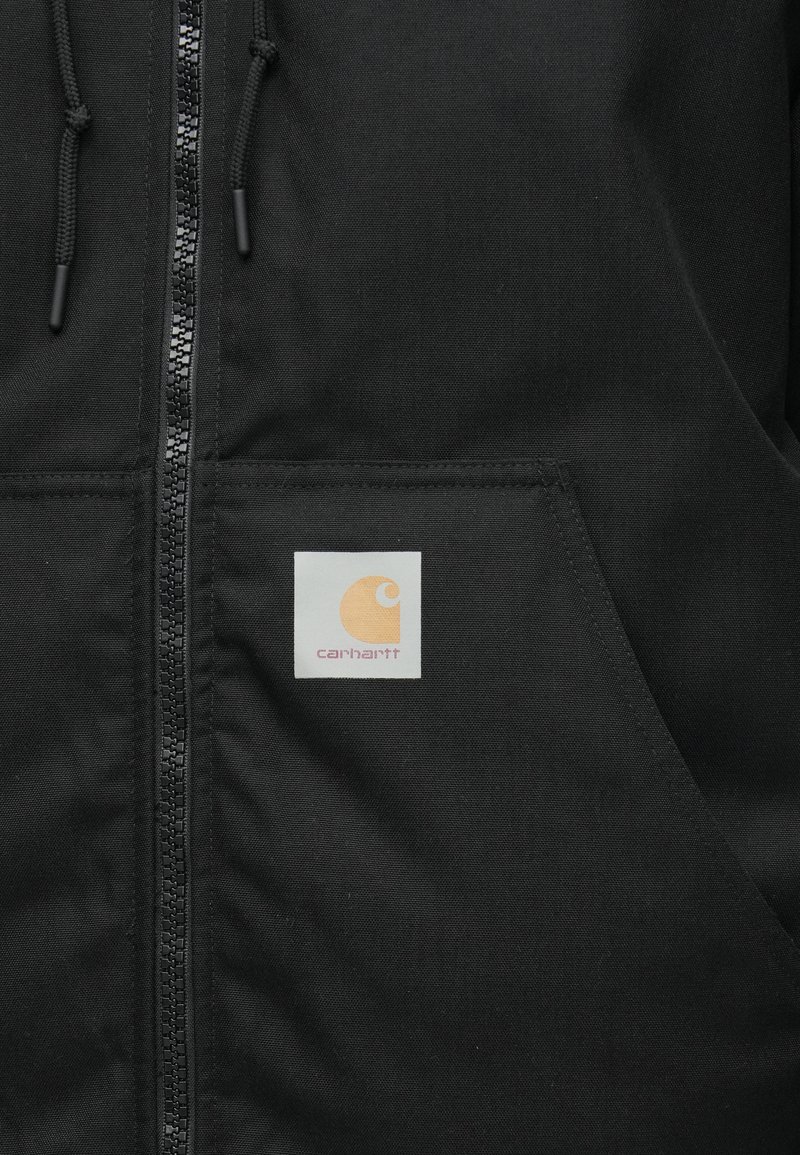 Chaqueta negra con cremallera frontal, puños acanalados y un parche con el logo de Carhartt en naranja y crema. Cuenta con una textura de tela suave y bolsillos.