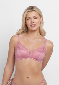 Blondinka v roza modrčku z vezenimi čipkami in ujemajočem se spodnjem perilu, ki se postavlja pred preprostim svetlo sivim ozadjem.