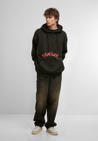 Sudadera negra con un gráfico rojo de "THUG LIFE" en la parte frontal, combinada con jeans oscuros de corte holgado y zapatillas beige. Capucha y bolsillo tipo canguro.