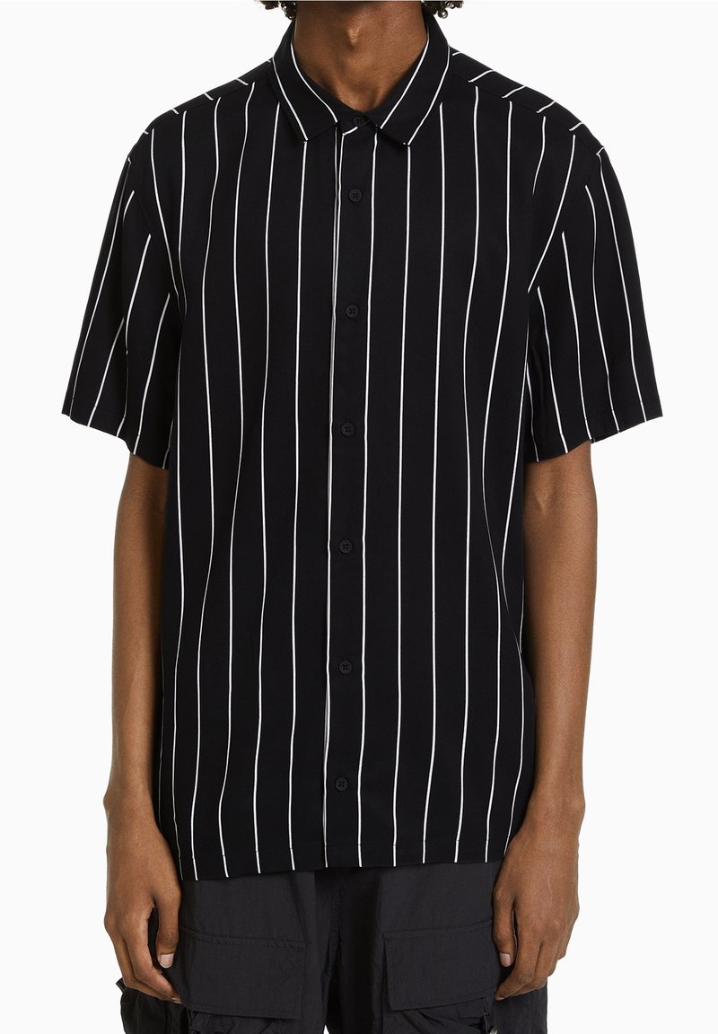 Chemise noire à manches courtes avec des rayures verticales blanches portée par une personne à la peau foncée et aux cheveux bouclés, montrée des épaules jusqu’aux hanches.