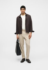 Homme debout portant une veste marron foncé, une chemise blanche, un pantalon beige, des chaussures marron foncé, tenant un grand sac cabas noir devant un fond uni.