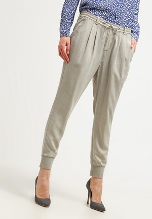 Mujer vistiendo pantalones jogger ajustados de color gris claro con cintura ajustable y puños acanalados, combinados con tacones altos gris oscuro y blusa azul con estampado.
