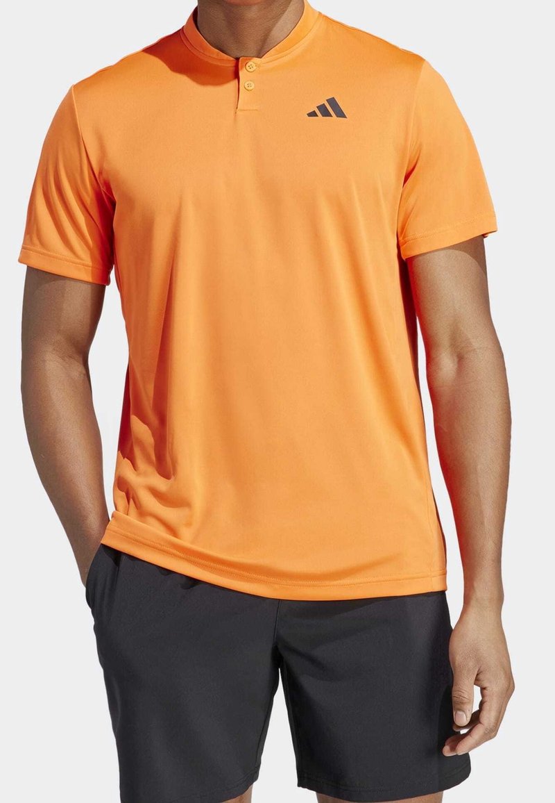 Homme portant une chemise à manches courtes orange vif avec deux boutons et un short noir, debout avec une main dans la poche.