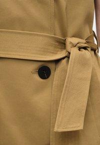 Ζώνη από ταμπά ύφασμα δεμένη με κόμπο πάνω σε ταιριαστό ένδυμα με εμφανές μαύρο κουμπί και ραφή για κουμπότρυπα.