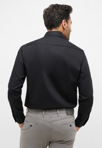Zwarte lange mouwen shirt met een klassieke kraag, gladde textuur en met knopen afsluitbare manchetten, gedragen met lichte grijze broek met achterzakken.