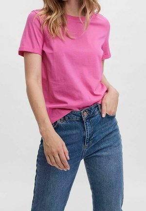 Vrouw draagt een felroze kortärmelig T-shirt, lichtjes ingestopt in een blauw high-waisted jeans met één hand in de zak.
