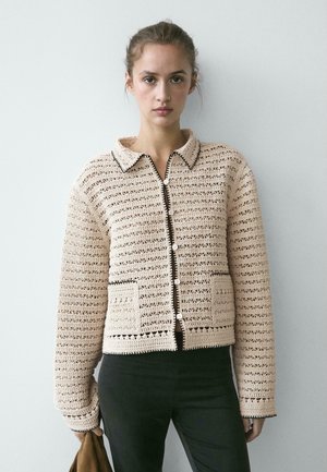 Giovane donna che indossa una giacca beige all'uncinetto con bottoni, rifinita in nero e con due tasche frontali, che tiene un oggetto marrone nella mano sinistra.