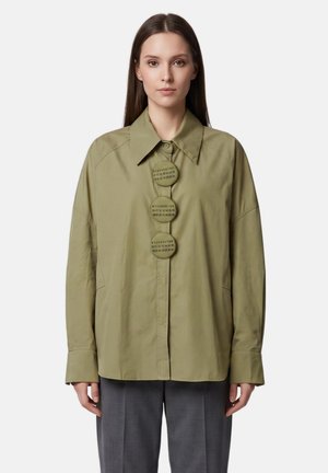 Donna che indossa una camicia oversize verde oliva con grandi bottoni rotondi con numeri stampati, abbinata a pantaloni grigi.