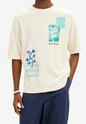 Mężczyzna w kremowej koszuli z krótkim rękawem, ozdobionej niebieskimi kwiatowymi i falistymi naszywkami, w połączeniu z granatowymi spodniami i białą bransoletką.