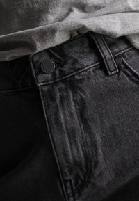 Jean en denim noir avec une surface texturée, dotés d'une fermeture à bouton métallique et d'une coupe droite, partiellement recouverts par une chemise grise.