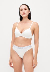 Soutien-gorge blanc avec un décolleté en V profond et de fines bretelles, accompagné d'un string blanc avec une ceinture "HUGO" grise. Texture lisse, design minimal.
