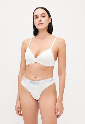 Witte beha met een diepe V-hals en dunne bandjes, gecombineerd met witte string-onderbroek met een grijze "HUGO" tailleband. Glad textuur, minimale uitstraling.