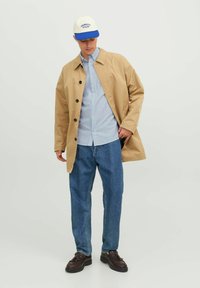 Helle beige Knopfjacke mit Kragen, kombiniert mit einem blau-weißen gestreiften Hemd, blauen Jeans und dunkelbraunen Schuhen.