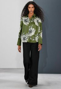 Zelená kvetinová košeľa na gombíky s bielymi kvetinovými vzormi, kombinovaná s čiernymi širokými nohavicami. Viditeľná je hladká látka a dlhé rukávy.
