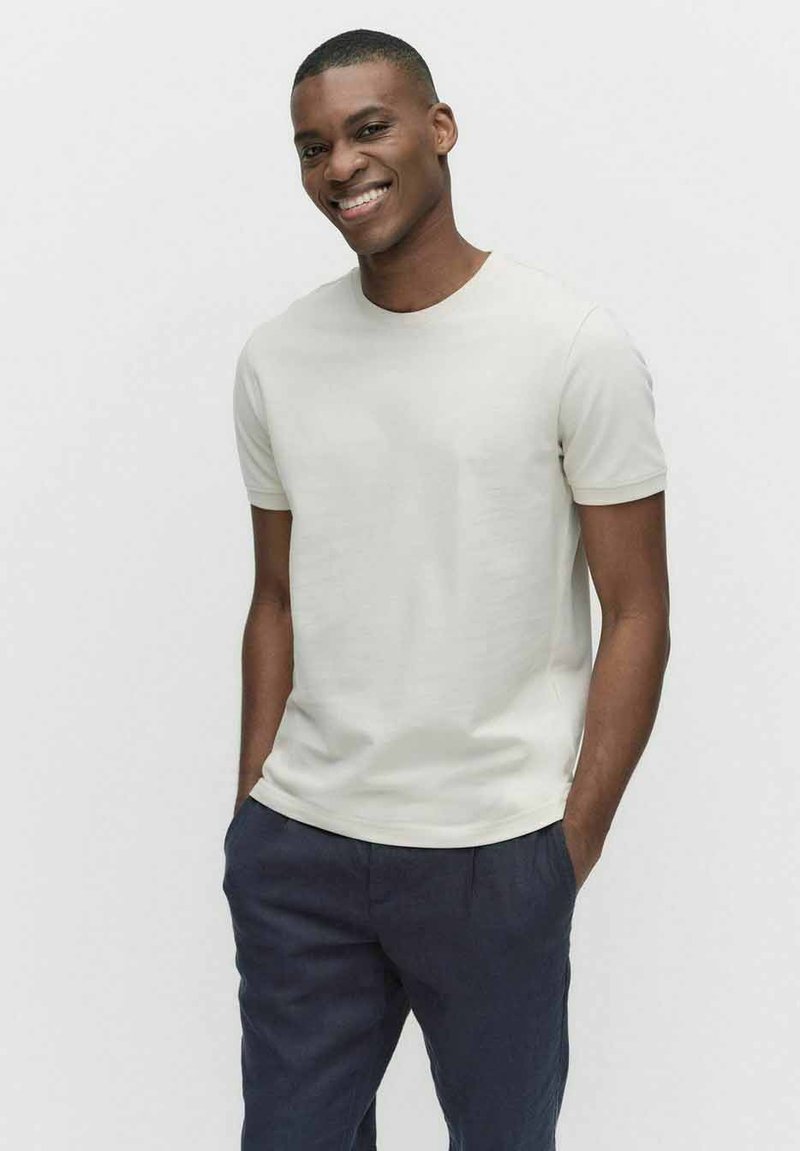 Ljusbeige t-shirt med kort ärm, tillverkad av mjuk bomull, med rund halsringning och en figurnära silhuett, tillsammans med marinblå byxor.