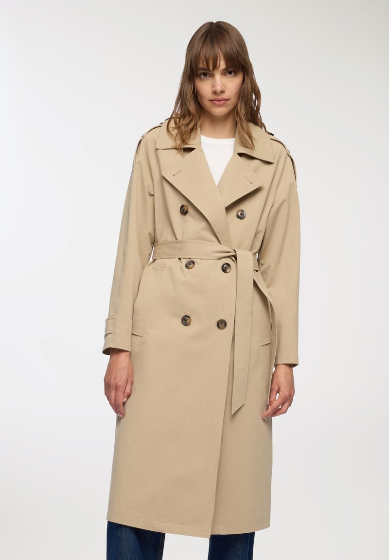 Beżowy płaszcz trench z podwójnym zapięciem, dużymi guzikami i przewiązaniem w talii. Oferuje szeroki kołnierz i długie rękawy. Gładka tekstura materiału.
