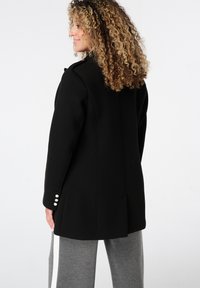 Zwarte op maat gemaakte jas met een gestructureerd ontwerp, voorzien van een enkele rij zilveren knopen op de manchetten en een strakke, moderne silhouet.