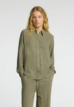 Camicia a maniche lunghe color verde oliva, con bottoni, realizzata in un tessuto leggero con una texture liscia. Presenta un orlo curvo e due tasche laterali.