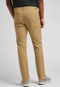 Beige farkut, joissa on suora leikkaus ja joissa on kaksi takataskua kangasdetaililla, yhdistettynä harmaisiin urheilujalkineisiin.