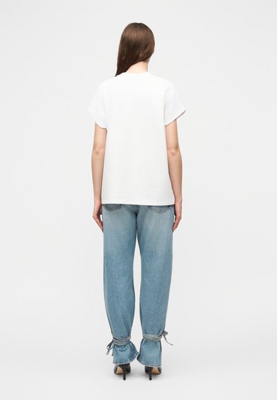 Γυναίκα με μακριά καστανά μαλλιά, φοράει λευκό κοντομάνικο πουκάμισο, ανοιχτό μπλε τζιν με δεμένα μανίκια και μαύρα ψηλοτάκουνα παπούτσια, στέκεται γυρισμένη προς τα πίσω.