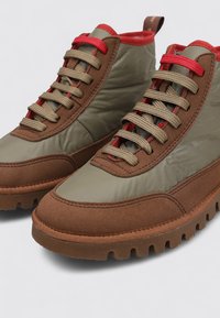Zapatillas altas marrones y verdes con parte superior de ante texturizado y nailon, que cuentan con acentos y cordones rojos, además de una gruesa suela de goma con tacos.