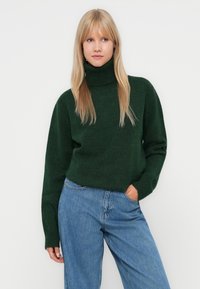 Pull à col roulé vert en matière douce, avec une coupe décontractée, des poignets côtelés, porté avec un jean bleu clair.