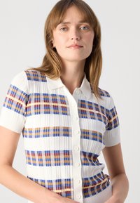 Jeune femme aux cheveux châtain clair portant une chemise blanche à manches courtes avec des motifs en plaid bleu, orange et rouge.