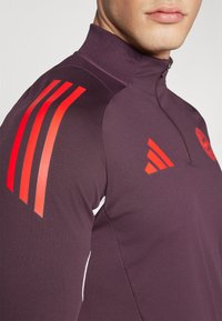 Giacca a vento imbottita con colletto alto in tessuto viola scuro, caratterizzata da strisce di accento rosse sulle spalle e un piccolo logo sul petto.