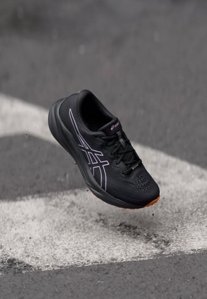 Schwarzer Asics-Laufschuh mit oranger Sohle, der in der Luft über strukturiertem Asphalt mit weißen Straßenmarkierungen aufgenommen wurde.