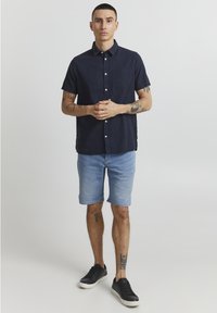 Camisa de botones de manga corta en color marino, pantalones cortos de mezclilla azul claro, zapatillas negras, con un bolsillo en la camisa y tatuajes visibles en los brazos.