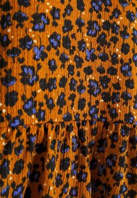 Volangklänning i orange tyg med en texturerad yta. Har svarta och lila blommönster mot en livlig bakgrund.